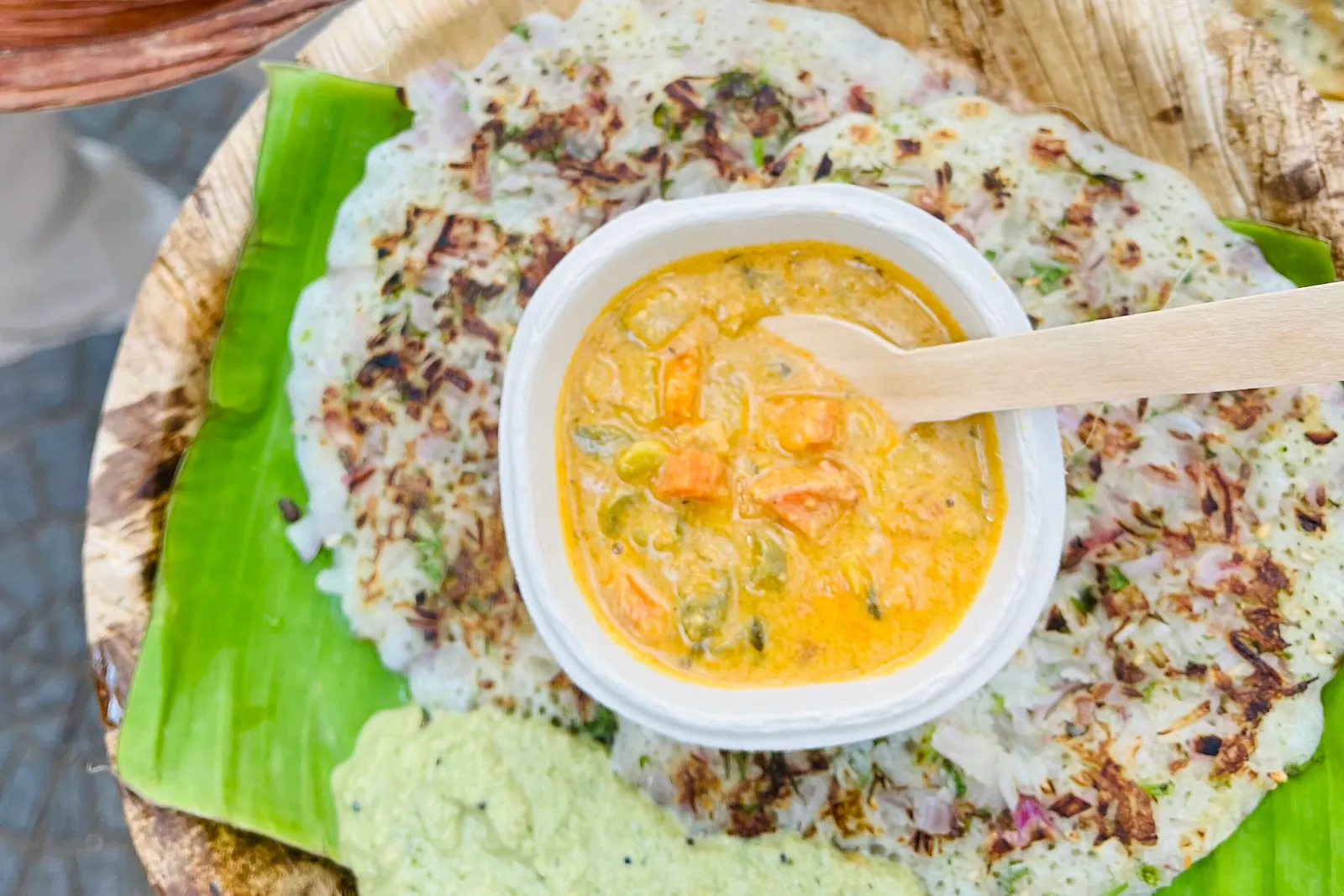 An onion uttapam served on a banana leaf in a palm-leaf plate at The Rameshwaram Cafe, with a cup of sambar in the middle and coconut chutney alongside