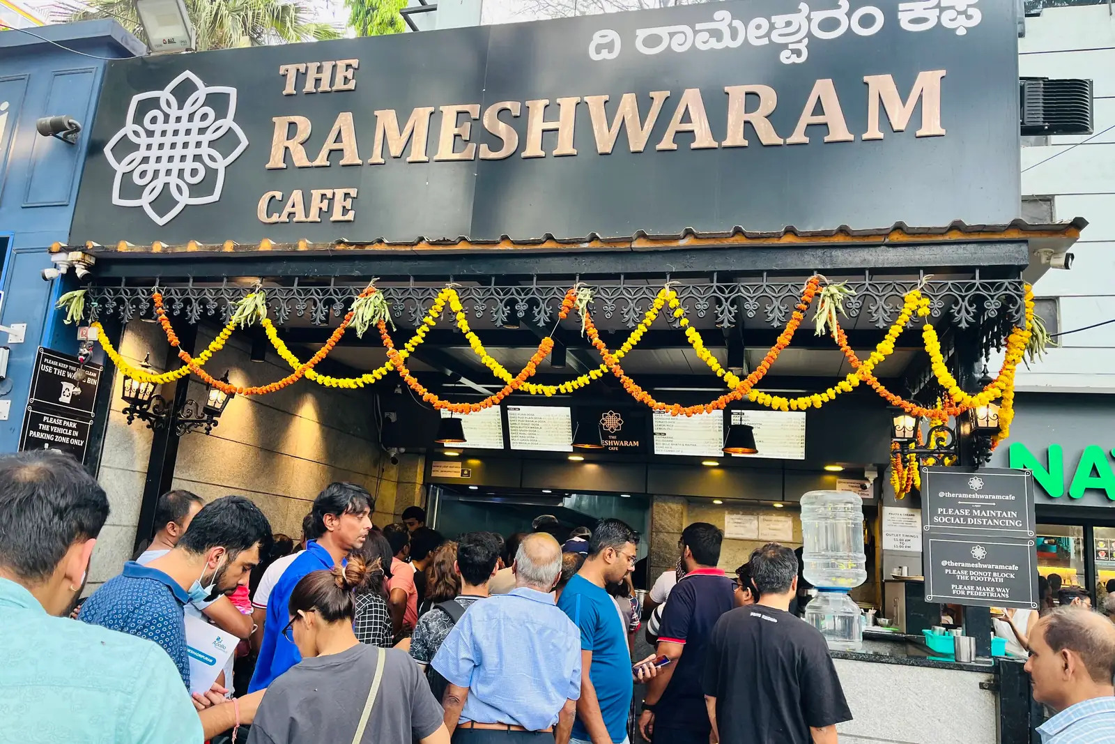 A crowd pressed against the ordering counter at The Rameshwaram Cafe in Bengaluru, marigold garlands strung across the frontage above them
